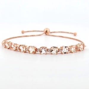 May include: A rose gold adjustable bracelet featuring a series of square and round-cut peach-colored gemstones. Small clear accent stones are interspersed between the larger gems. The bracelet is displayed against a white background.