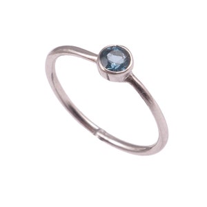 Op de afbeelding: Een delicate zilveren ring met een ronde, lichtblauwe edelsteen. De ring heeft een dunne band en een eenvoudig, elegant ontwerp. De edelsteen is gezet in een bezel setting, tegen een witte achtergrond.
