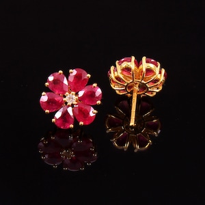 Könnte beinhalten: Ein Paar goldene Ohrringe mit einem Blumenmuster. Die Ohrringe sind aus roten Rubinen und einem einzelnen Diamanten in der Mitte jeder Blume gefertigt.