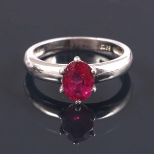 Peut inclure: Une bague en argent ornée d'un grand rubis de forme ovale. Le rubis est d'un rouge vif et est serti dans une monture à six griffes. L'anneau de la bague est lisse et poli, reflétant la lumière. La bague est présentée sur une surface noire réfléchissante.