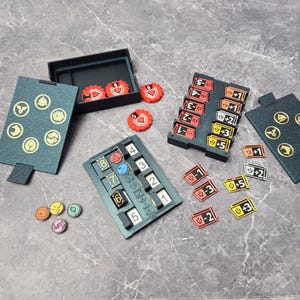 May include: A selection of game components, including dark teal game boards with gold symbols, a black box, red tokens, and various coloured tokens. There are also black and red tokens with numbers and symbols.