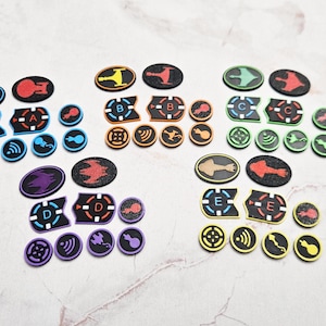 May include: A collection of colorful, die-cut game tokens with various shapes and designs. The tokens feature black, red, blue, green, yellow, and purple accents. The tokens have letters A, B, C, D, and E, and other symbols.