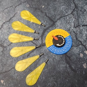 May include: A blue and yellow circular dial with a red pointer and numbers 1 through 12, labeled "am" and "pm".  The dial is surrounded by seven yellow, leaf-shaped objects with silver tips.