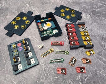 May include: A tabletop game accessory set featuring a black storage tray, two black lids with yellow circular icons, a black tracker board with numbered slots, and various colorful status tokens labeled with text like ASSAULT, STUNNED, GANKING, and EMPOWERED.