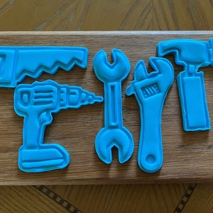 May include: Five blue plastic cookie cutters shaped like tools: a saw, a drill, a wrench, a wrench, and a hammer.