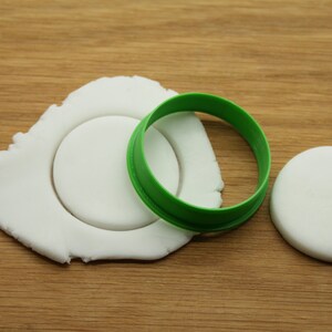 May include: A white fondant circle being cut out with a green plastic cookie cutter. There is a second white fondant circle to the right of the cutter.