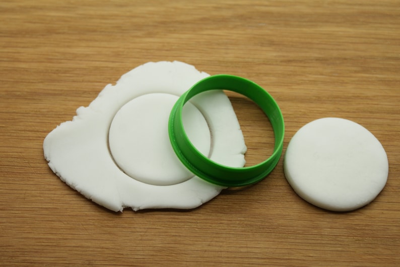 May include: A white fondant circle being cut out with a green plastic cookie cutter. There is a second white fondant circle to the right of the cutter.