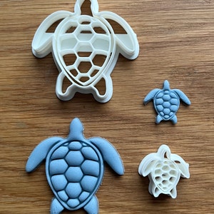 May include: Three turtle cookie cutters, two are white and one is blue. The largest cutter is white and has a detailed shell design. The smallest cutter is also white and has a simple shell design. The middle cutter is blue and has a detailed shell design.