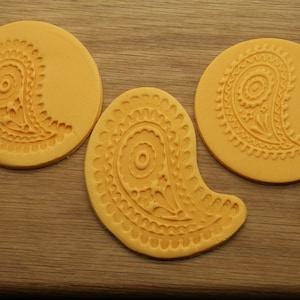 May include: Three yellow silicone moulds with a paisley design. The moulds are on a wooden surface.