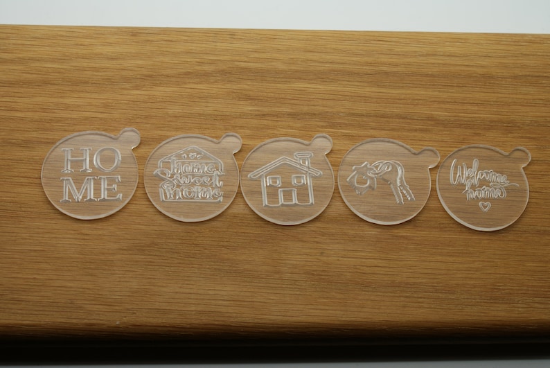 May include: Five clear plastic cookie cutters with different designs. The cutters are on a wooden surface. The designs include the words "Home", "Sweet Home", a house, a heart, and the words "Welcome Home".