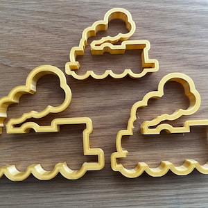 May include: Three gold-coloured metal cookie cutters shaped like trains with smoke stacks. The cutters are different sizes.