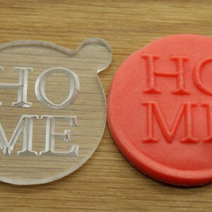 May include: A clear plastic cookie cutter and a red cookie with the word "HOME" embossed on it.
