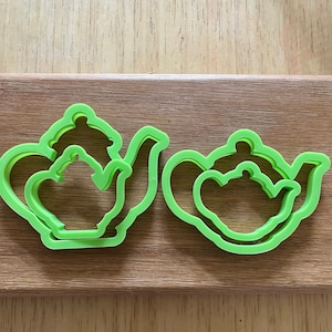 May include: Two green plastic cookie cutters shaped like teapots. The cutters are layered, with a smaller teapot inside a larger teapot.