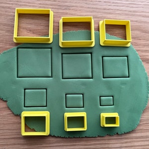 May include: A set of nine yellow plastic cookie cutters in various sizes and shapes, arranged on a green surface. The cutters are shaped like rectangles and squares.