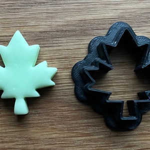 May include: A green maple leaf cookie cutter and a green maple leaf cookie. The cookie cutter is made of black plastic.
