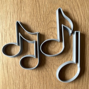 May include: Three grey plastic cookie cutters shaped like musical notes. The cutters are on a wooden surface.