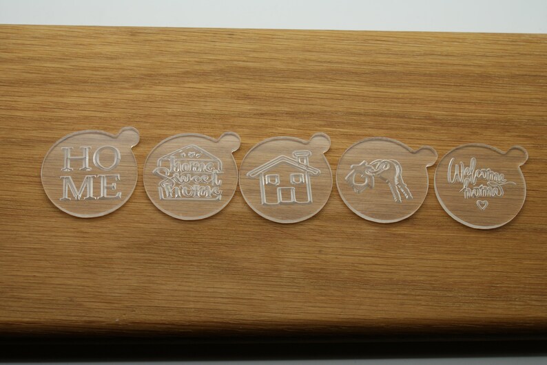 May include: Five clear plastic cookie cutters with different designs. The cutters are on a wooden surface. The designs include the words "Home", "Sweet Home", a house, a heart, and the words "Welcome Home".