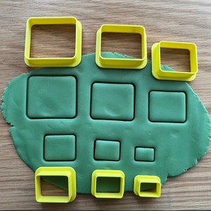 May include: A set of nine yellow plastic cookie cutters in various sizes, arranged on a green surface. The cutters are shaped like squares and rectangles.