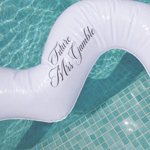 May include: A white inflatable pool float with a curved, elongated shape. The surface features the black text "Future Mrs Gamble" written in a cursive script font. The float is positioned over clear blue water with a tiled pool floor visible.