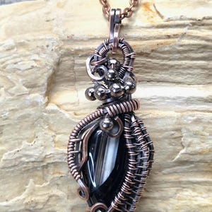 May include: A handcrafted pendant necklace featuring a black and white stone encased in copper wire. The wire is intricately woven and wrapped around the stone, with small silver-colored beads adding detail. The pendant hangs from a copper-colored chain.