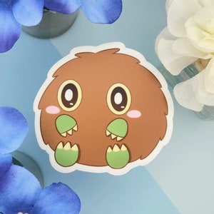 May include: A die-cut sticker of a cartoon creature with a brown, fluffy body, large oval eyes, and small green feet. The creature has pink cheeks. The background is light blue with floral accents.