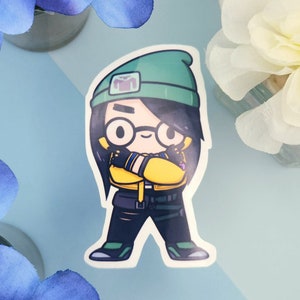 Könnte beinhalten: Ein Aufkleber mit einer Zeichentrickfigur mit schwarzen Haaren, Brille und grüner Mütze. Die Figur trägt eine gelbe Jacke, eine schwarze Hose und grüne Schuhe. Die Figur hat die Arme verschränkt. Der Aufkleber befindet sich auf einer hellblauen Oberfläche.