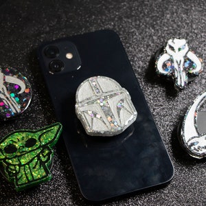 May include: Five phone grips with different designs. The grips are made of resin and have glitter embedded in them. The designs include a Mandalorian helmet, a Baby Yoda, and three different versions of the Mandalorian symbol.