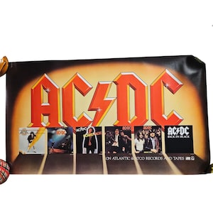 Op de afbeelding: Een vintage AC/DC poster met het bandlogo in oranje en een gouden achtergrond. De poster toont albumhoezen waaronder "Highway to Hell" en "Back in Black". De tekst onderaan luidt "ON ATLANTIC & ATCO RECORDS AND TAPES".