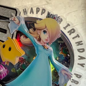 May include: A layered cake topper featuring a character with blonde hair in a light blue dress and a silver crown, accompanied by a yellow star figure. The circular frame contains colorful confetti and the text HAPPY BIRTHDAY printed in silver glitter.
