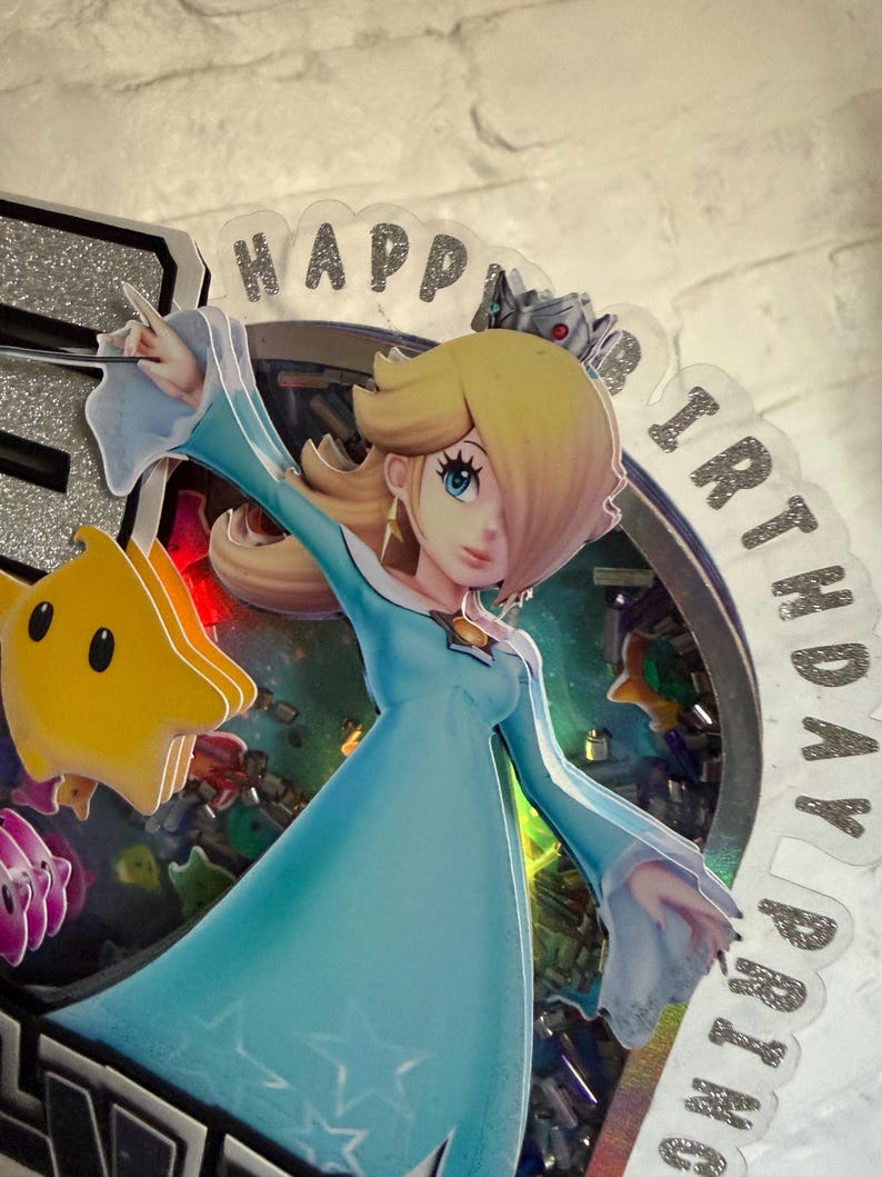May include: A layered cake topper featuring a character with blonde hair in a light blue dress and a silver crown, accompanied by a yellow star figure. The circular frame contains colorful confetti and the text HAPPY BIRTHDAY printed in silver glitter.