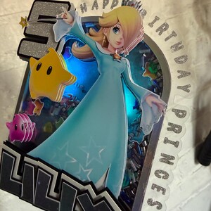 May include: A custom cake topper featuring a layered design with the character Rosalina in a light blue dress, a yellow star, a pink star, and the number 5. The text reads HAPPY BIRTHDAY PRINCESS and LILLY. It includes blue LED lighting.