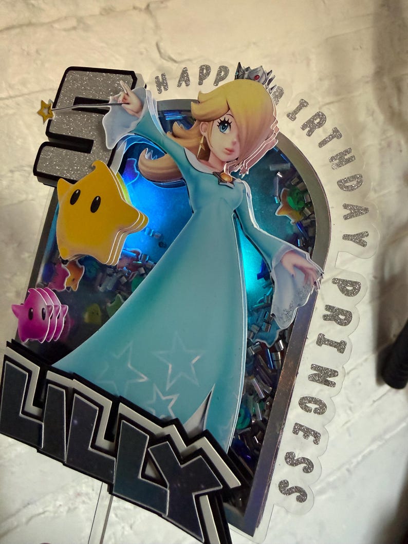 May include: A custom cake topper featuring a layered design with the character Rosalina in a light blue dress, a yellow star, a pink star, and the number 5. The text reads HAPPY BIRTHDAY PRINCESS and LILLY. It includes blue LED lighting.