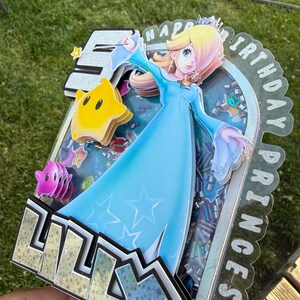May include: A layered cake topper featuring a cutout of Princess Rosalina in a blue dress, a yellow Luma, and the name LILLY in bold letters. The design includes a shaker element filled with colorful sequins and the text HAPPY BIRTHDAY PRINCESS.