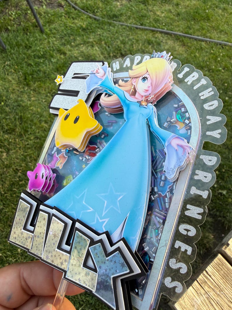 May include: A layered cake topper featuring a cutout of Princess Rosalina in a blue dress, a yellow Luma, and the name LILLY in bold letters. The design includes a shaker element filled with colorful sequins and the text HAPPY BIRTHDAY PRINCESS.