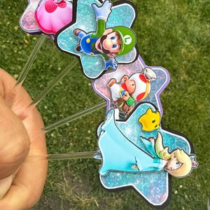 Puede incluir: Cuatro adornos para pasteles con personajes de Super Mario, incluyendo a la Princesa Peach, Luigi, Toad y Rosalina. Cada personaje est&aacute; montado sobre un recorte en forma de estrella con un acabado brillante azul o morado, unido a un palito de pl&aacute;stico.