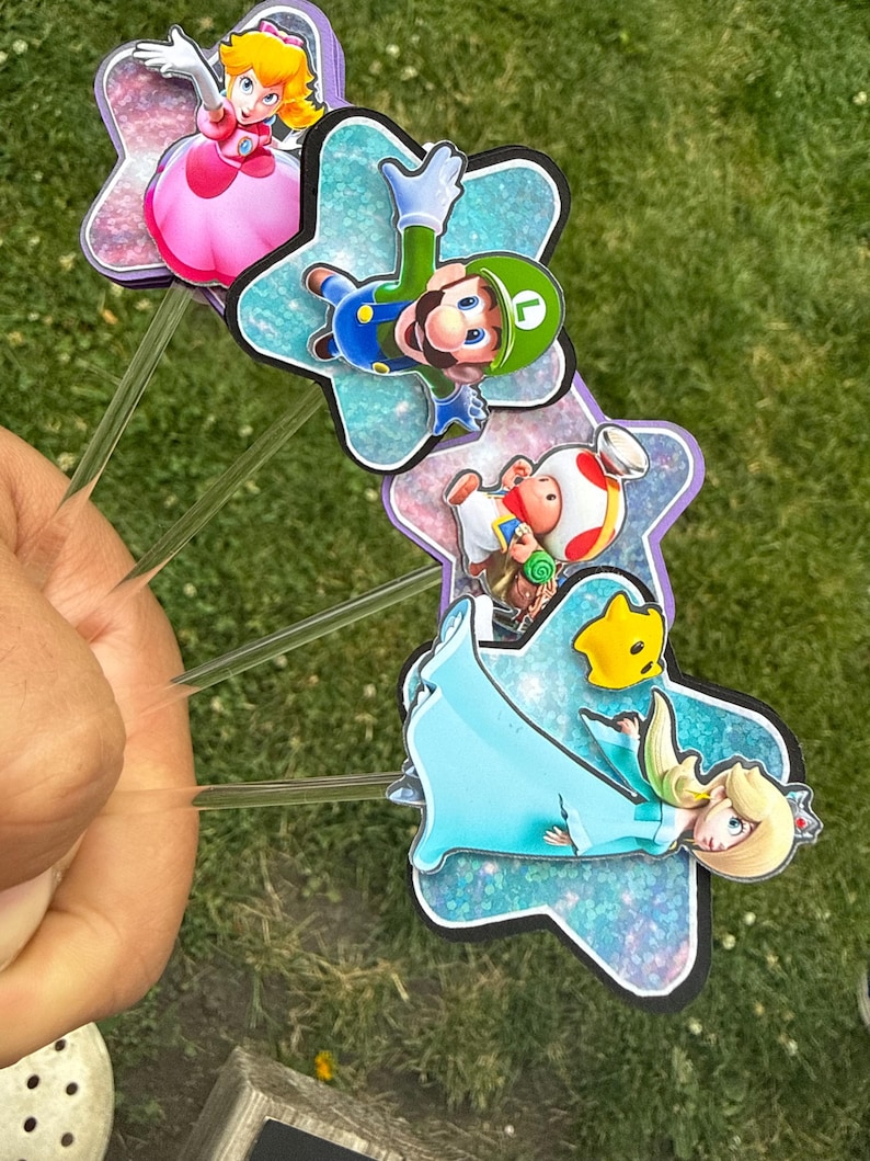 Puede incluir: Cuatro adornos para pasteles con personajes de Super Mario, incluyendo a la Princesa Peach, Luigi, Toad y Rosalina. Cada personaje est&aacute; montado sobre un recorte en forma de estrella con un acabado brillante azul o morado, unido a un palito de pl&aacute;stico.
