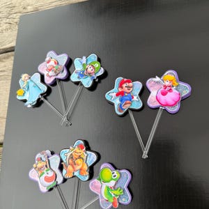 Puede incluir: Un conjunto de siete adornos para cupcakes con varios personajes de Super Mario, incluyendo a Mario, Luigi, Peach, Bowser y Yoshi. Cada personaje est&aacute; montado sobre una base en forma de estrella con purpurina unida a un palillo de pl&aacute;stico transparente.