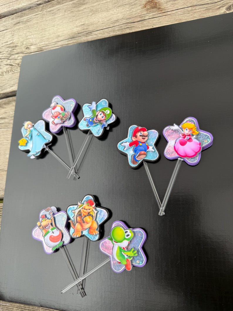 Puede incluir: Un conjunto de siete adornos para cupcakes con varios personajes de Super Mario, incluyendo a Mario, Luigi, Peach, Bowser y Yoshi. Cada personaje est&aacute; montado sobre una base en forma de estrella con purpurina unida a un palillo de pl&aacute;stico transparente.