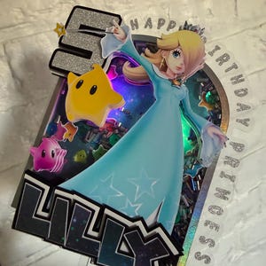 May include: A layered cake topper featuring the character Rosalina in a light blue dress, a yellow star, and the number 5 in silver glitter. The design includes the text HAPPY BIRTHDAY PRINCESS and the name LILLY in black, with colorful confetti.