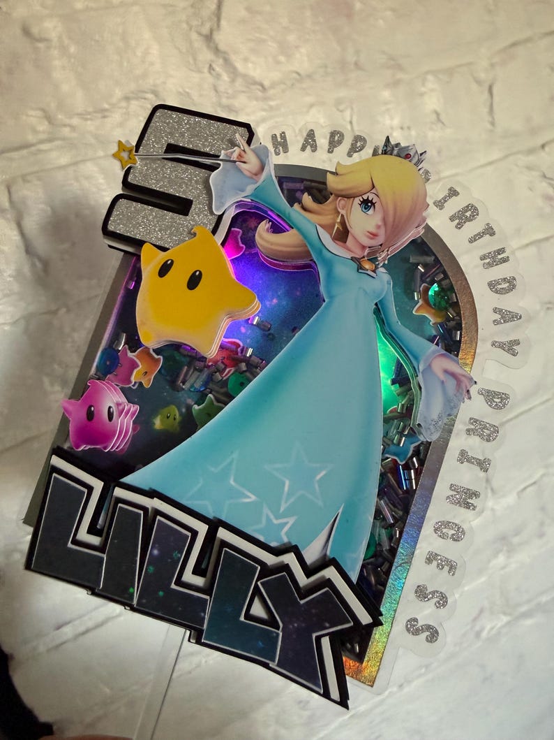 May include: A layered cake topper featuring the character Rosalina in a light blue dress, a yellow star, and the number 5 in silver glitter. The design includes the text HAPPY BIRTHDAY PRINCESS and the name LILLY in black, with colorful confetti.