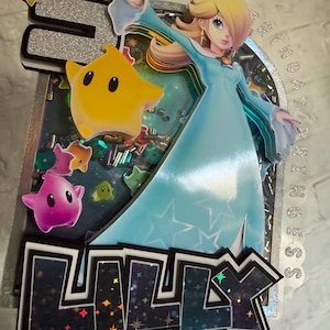 May include: A layered cake topper featuring a glittery silver number 5, a character in a light blue gown, and a yellow star. The name LILLY is displayed in dark, star-patterned letters at the bottom. The design includes colorful star-shaped cutouts.