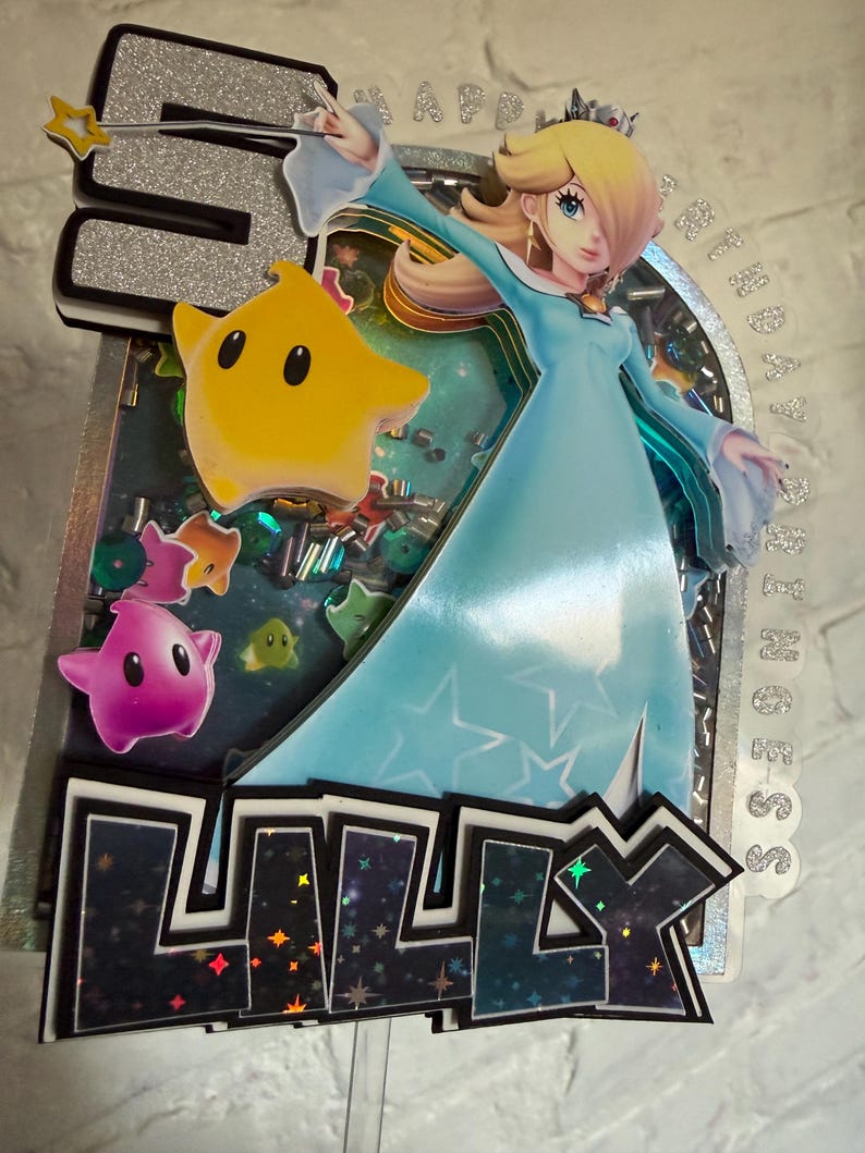 May include: A layered cake topper featuring a glittery silver number 5, a character in a light blue gown, and a yellow star. The name LILLY is displayed in dark, star-patterned letters at the bottom. The design includes colorful star-shaped cutouts.