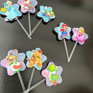 Puede incluir: Un conjunto de ocho adornos para pasteles con varios personajes de videojuegos, incluidos Mario, Luigi, Peach, Bowser y Toad. Cada personaje est&aacute; montado sobre un recorte en forma de estrella con acabado brillante y un palito de pl&aacute;stico transparente.