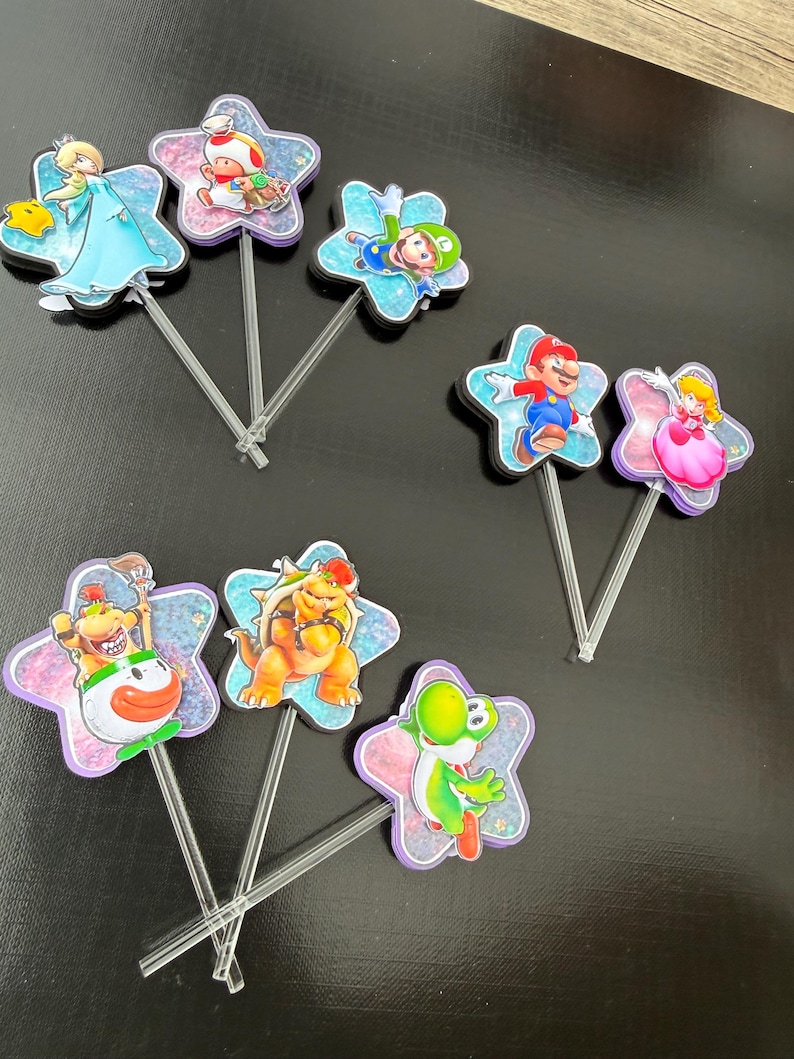 Puede incluir: Un conjunto de ocho adornos para pasteles con varios personajes de videojuegos, incluidos Mario, Luigi, Peach, Bowser y Toad. Cada personaje est&aacute; montado sobre un recorte en forma de estrella con acabado brillante y un palito de pl&aacute;stico transparente.