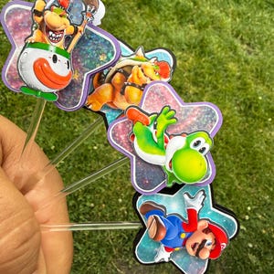 Puede incluir: Un conjunto de cuatro adornos para cupcakes con coloridos personajes de Nintendo, incluyendo a Bowser Jr. en un coche payaso, Bowser, Yoshi y Mario. Cada personaje est&aacute; montado sobre un recorte en forma de estrella con acabado hologr&aacute;fico.