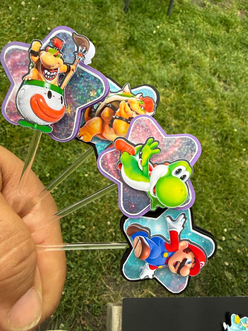 Puede incluir: Un conjunto de cuatro adornos para cupcakes con coloridos personajes de Nintendo, incluyendo a Bowser Jr. en un coche payaso, Bowser, Yoshi y Mario. Cada personaje est&aacute; montado sobre un recorte en forma de estrella con acabado hologr&aacute;fico.