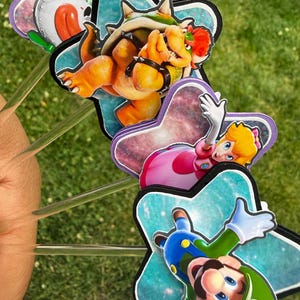 Puede incluir: Un juego de cuatro adornos decorativos para pasteles con ilustraciones coloridas de Bowser, la Princesa Peach y Luigi sobre fondos en forma de estrella. Cada figura est&aacute; montada en un palito de pl&aacute;stico transparente para pasteles.