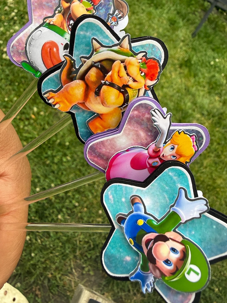 Puede incluir: Un juego de cuatro adornos decorativos para pasteles con ilustraciones coloridas de Bowser, la Princesa Peach y Luigi sobre fondos en forma de estrella. Cada figura est&aacute; montada en un palito de pl&aacute;stico transparente para pasteles.