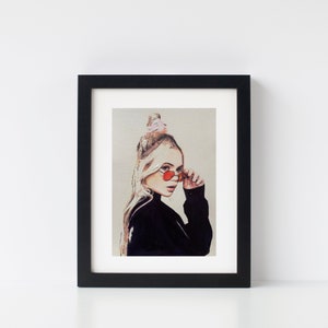 May include: A framed print of a painting depicting a woman with blonde hair wearing red heart-shaped sunglasses and a black turtleneck sweater. The woman is looking to the side and has a pink hair tie in her hair.