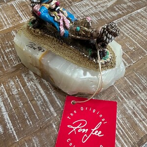 May include: A decorative figurine of a clown lying on a bed of gold beads, atop a white stone base. The clown wears a blue jacket, pink scarf, and a red nose. A red tag reads "The Original Ron Lee Collection."